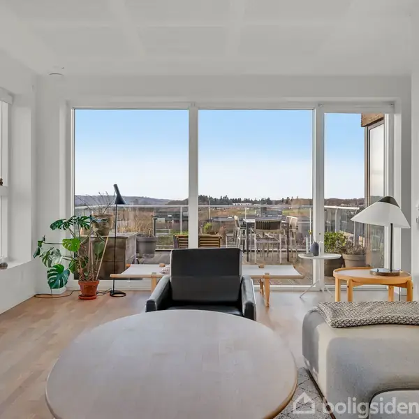 En moderne stue med sorte og grå møbler står stilfuldt mod store panoramavinduer, der giver udsigt til en terrasse og et landskab i baggrunden. Trægulv og minimalistisk indretning skaber rolig atmosfære.