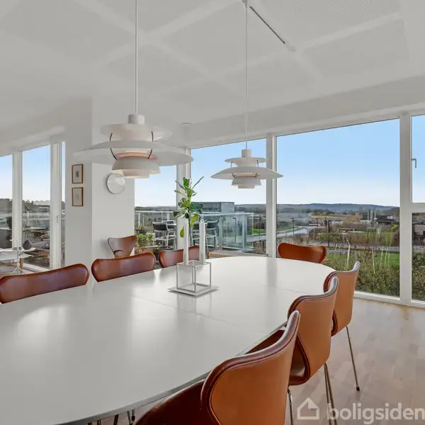 Et stort, hvidt ovalt spisebord med brune stole står i et lyst rum med store vinduer, der giver udsigt over et landskab. En elegant lysekrone hænger over bordet.