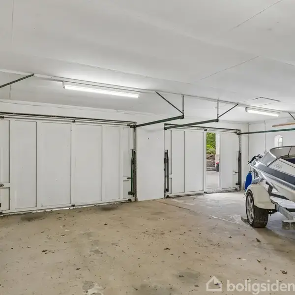 Båd på trailer står i garage med hvide vægge og gulv. Garageporte er åbnet. På venstre væg hænger haveredskaber og en stige. Garagen er rummelig og oplyst.