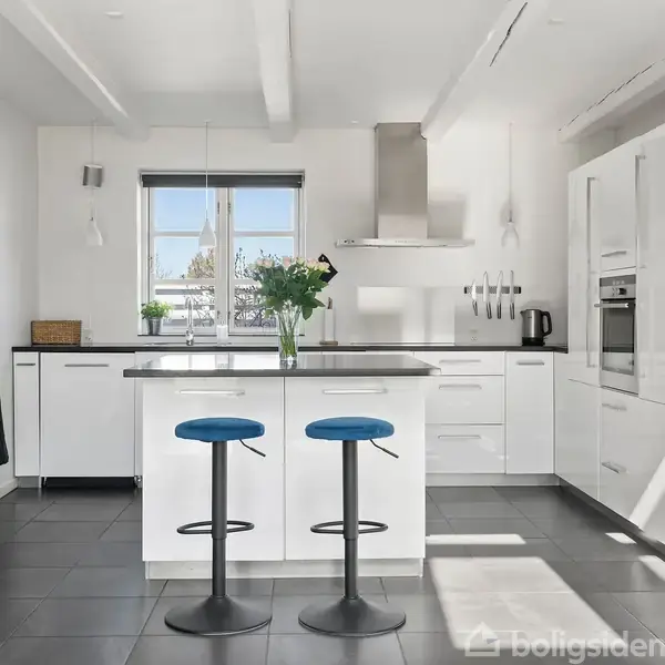 Moderne hvidt køkken med ø, hvor to blå barstole står. Vasen med blomster pynter øen. Køkkenmaskiner og skabsvæg omkranser rummet. Sollys strømmer ind gennem vinduet.