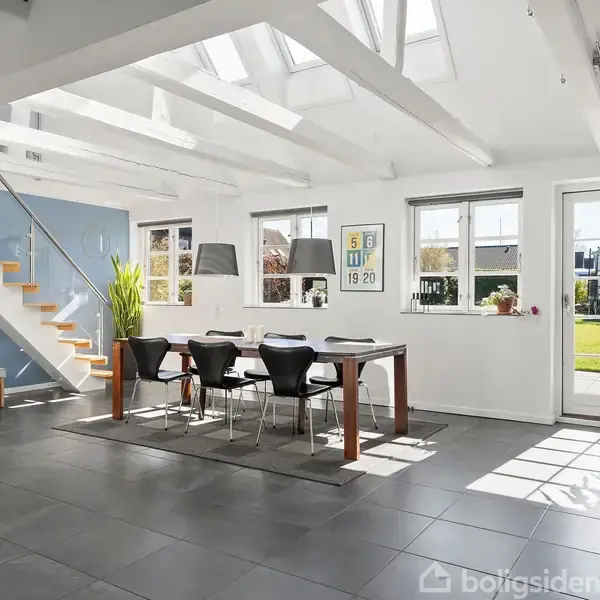 Et spisebord med sorte stole står i en lys stue med skiferfliser. Trappen til venstre fører til en overetage, og store vinduer giver udsigt til en have.