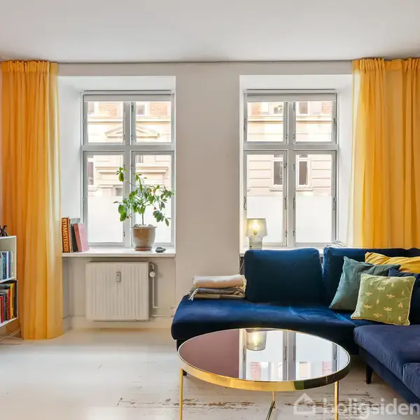 Blå sofa med puder er placeret i en stue med orange væg og gule gardiner, foran vinduer med lys. Rundt sofabord i midten, reol med bøger står mod væggen.
