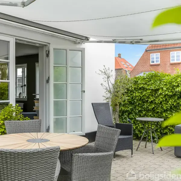 Terrassemøbler står på en flisebelagt terrasse foran en åben glasdør. Omkring er en tæt hæk, der afskærmer området. En markise er trukket ud over terrassen.