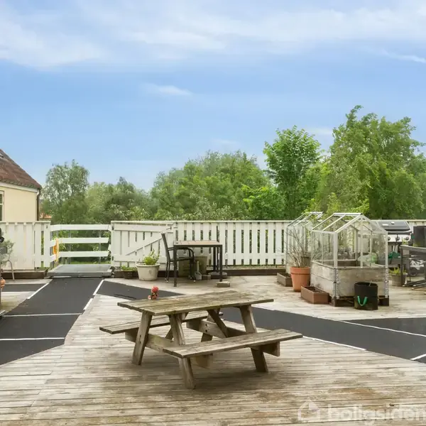 Et træbord står på en terrasse omgivet af planter, to drivhuse og en grill. Terrassen er omkranset af et hvidt stakit, og der er grønne træer i baggrunden under en blå himmel.
