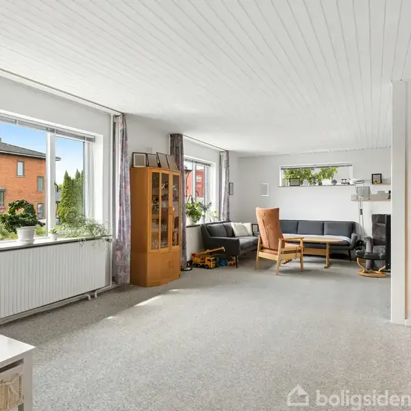 Stue med lysindfald fra store vinduer, sofagruppe og vitrineskab. Væggene er lyse, og der er dekorationer på reolerne. Udenfor ses et rødt murstenshus og grøn beplantning.