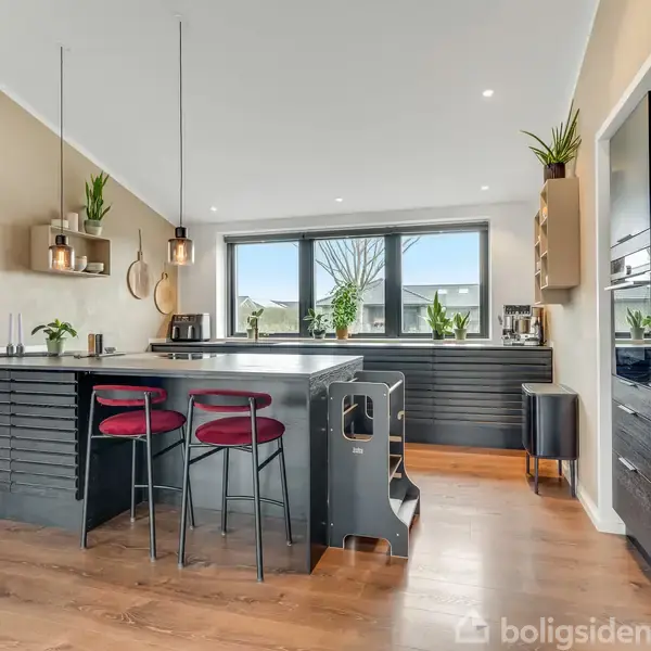 Køkken med sort køkkenø, tre barstole, og planter. Store vinduer giver lys. Moderne apparater indbygget i væggen. Maleri hænger på væggen. Trægulv skaber en varm atmosfære.