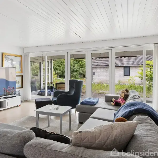 Stue med grå sofa og lænestol, hvor et barn sidder med en tablet. Store vinduer giver udsigt til en have. Der er en stor fladskærm og malerier på væggene.