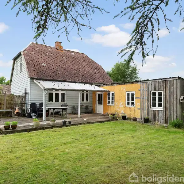 Et hus med en træterrasse og to udendørsstole står i en have med en grøn græsplæne. En udendørs pejs ses til venstre.