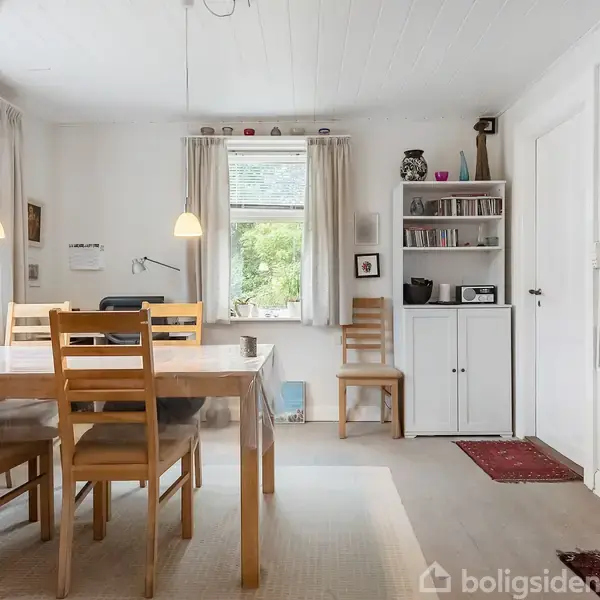 Spisebord med seks træstole i en lys spisestue. Hængelamper over bordet. En bogreol og stol ved vindue med udsigt til grønne træer. Forskelligt dekoration på hylderne.