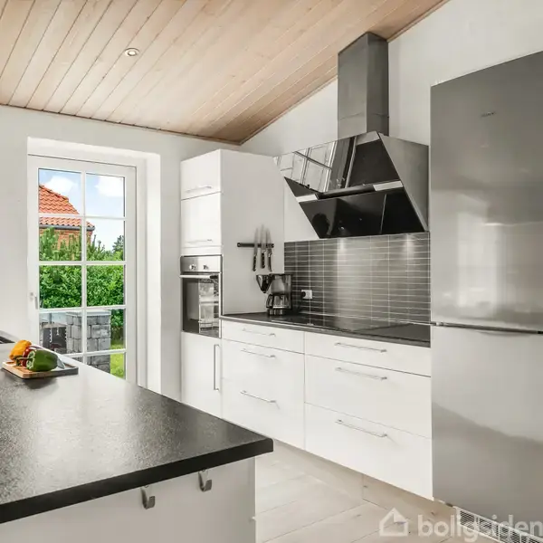 Et moderne køkken med hvide skabe og en stålgrå køleskabsvæg. En køkkenø med sort bordplade har frugter på toppen. Bagved ses en dør ud til en have med grønne planter.