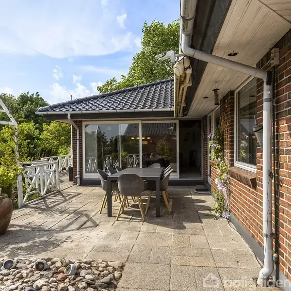 Hus med røde mursten og sort tag, udstyret med bord og stole på en terrasse. Omkranset af grønne træer og buske, med klinkebelagt underlag og udsigt til en have.
