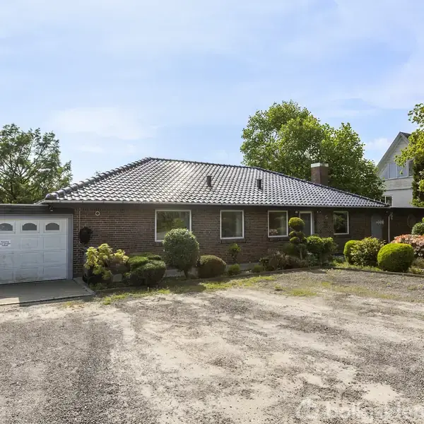 Et lavt, murstenshus med mørkt tag står foran en grusbelagt indkørsel. Huset har flere vinduer og en tilknyttet garage til venstre. Omgivet af velplejede buske og træer.