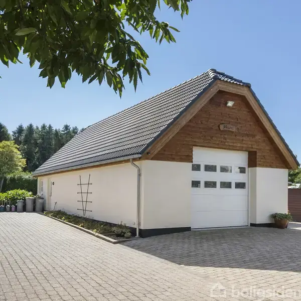 En hvid garagebygning med skråt tag står stille. Omgivet af en flisebelagt indkørsel og grøn beplantning. Solrigt og klart vejr under en blå himmel.