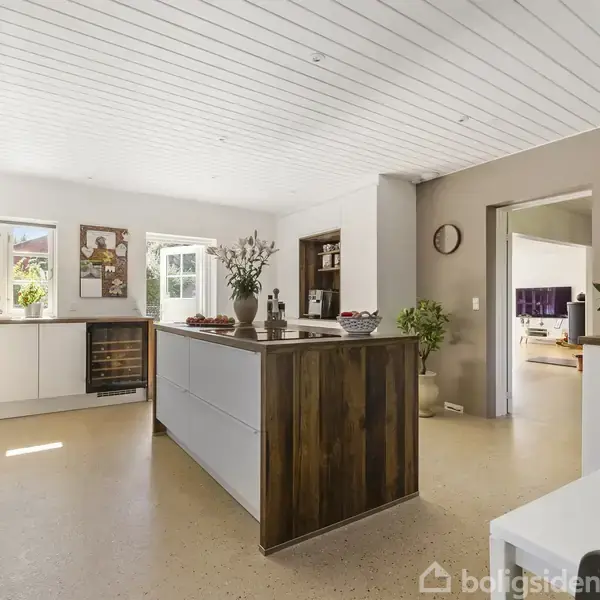 Et moderne køkken med en central køkkenø dekoreret med blomster. Hvide skabe omgiver rummet, oplyst af dagslys fra vinduerne. En åben forbindelse fører til en tilstødende stue.