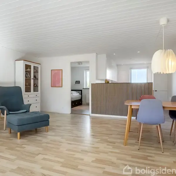 Stue med trægulv, hvor der står en blå lænestol og sofa til venstre. Et spisebord med farvede stole til højre. En høj lampe hænger over bordet.