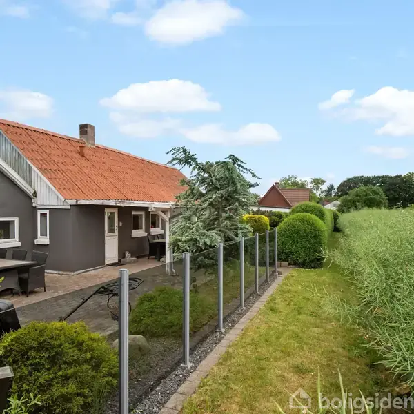 Gråt hus med rødt tegltag står ved siden af en glashegn og have. Bord og stole er på terrassen. Omkringliggende natur domineres af græs og buske under blå himmel.