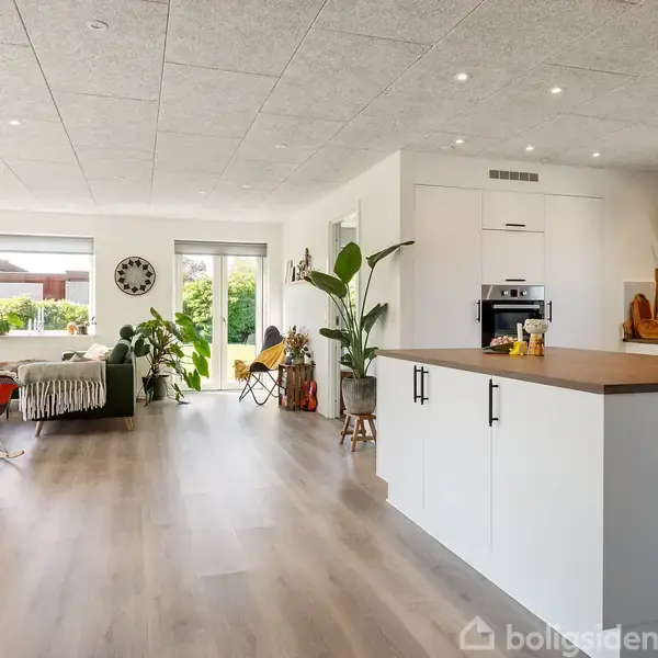 Et stort, lyst køkken-alrum med hvide skabe dominerer. Der ses en stue med sofa og planter i baggrunden. Store vinduer lukker naturligt lys ind.