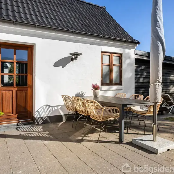 Havemøbler står på en flisebelagt terrasse foran et hvidt hus med en brun dør og vindue. Ved siden af bordet er en lukket parasol. Græsplæne ses i baggrunden.