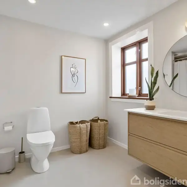 Et moderne badeværelse med et toilet, en stor, rund spejl over en trævask, og to flettede kurve ved siden af. Der hænger et abstrakt kunstværk på væggen.
