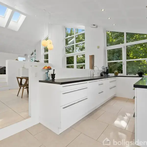 Et moderne køkken-alrum med højt til loftet. Et stort spisebord med stole står under runde lamper. Store vinduer giver udsigt til grønne træer. En glasdør fører ud til haven.