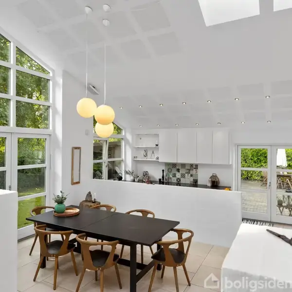 En moderne spisestue med sort bord og seks stole, beliggende i et lyst rum med store vinduer og hvidt køkken i baggrunden. Grøn udsigt gennem vinduerne.