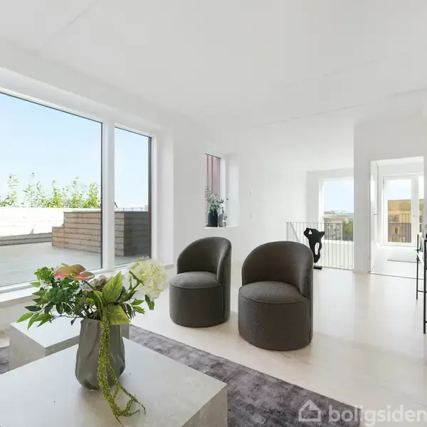 Stue med to mørke lænestole står på et lyst gulv ved siden af et sofabord med blomster. Store vinduer slipper dagslys ind, og der er udsigt til en terrasse.