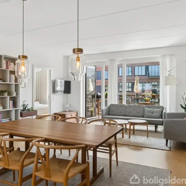 Spisebord med seks stole står i en lys stue. En sofa og sofabord ses foran store vinduer, der giver udsigt til bygninger udenfor. Bogreol til venstre.
