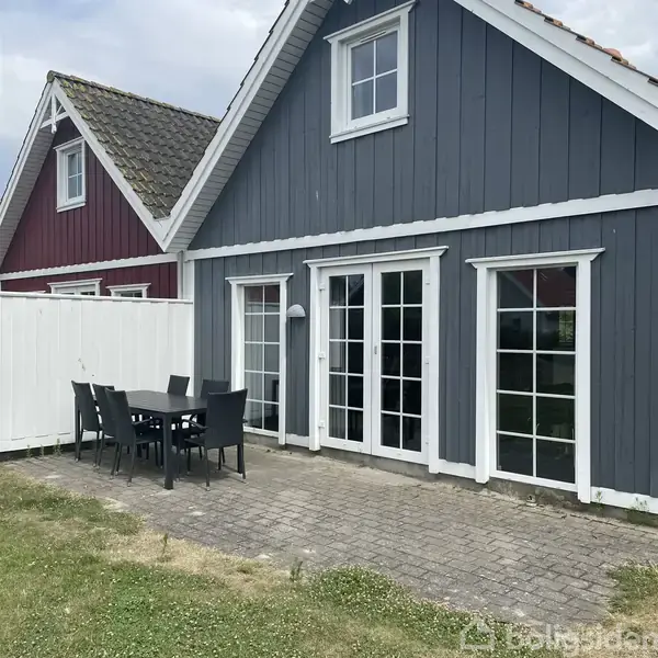 Gråmalet træhus med hvide vinduer og døre, bord og stole på en terrasse af stenfliser. Ved siden af det grå hus står en rød bygning. Græsplæne omslutter området.