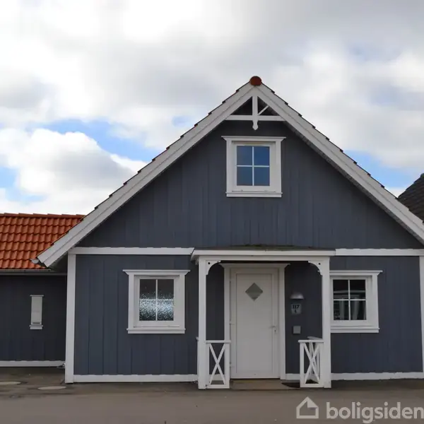 Gråmalet hus med skråtag, hvid rammedør og vinduer, står på flisebelagt grund. Venstre side forbinder til anden bygning med rødt tag. Skyet himmel i baggrunden.