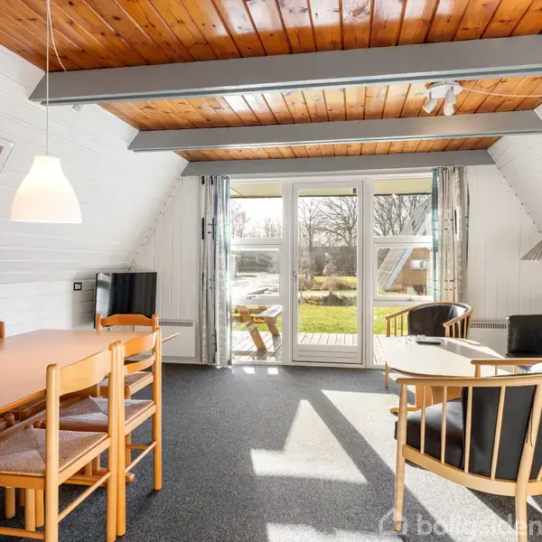 Et stueområde med spisebord og stole i træ, set i et hus med trægulv og loft. Et glasdør-område fører til en have, og to farverige kunstværker hænger på væggene.