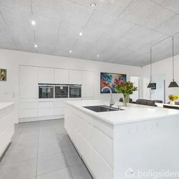 Et moderne køkken med hvide skabe og en ø i midten, indeholder en vask og blomster. Spisebordet med stole står i baggrunden, omgivet af kunst og hængende lamper.