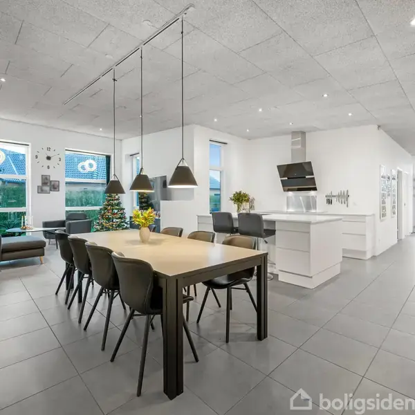 Et moderne køkken-alrum med et stort træbord omkranset af sorte stole, hængende lamper ovenover. I baggrunden ses en stue med sofa og juletræ ved vinduerne, og køkkenet har en central ø.