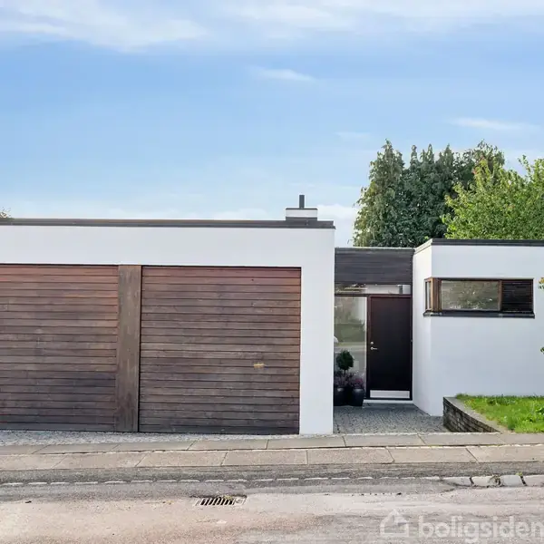 Et moderne hus med hvide vægge og trægarageporte. Skiltet ved hækken viser teksten "Til salg" og et firmanavn med kontaktnummer. Omgivet af grønne træer og have.