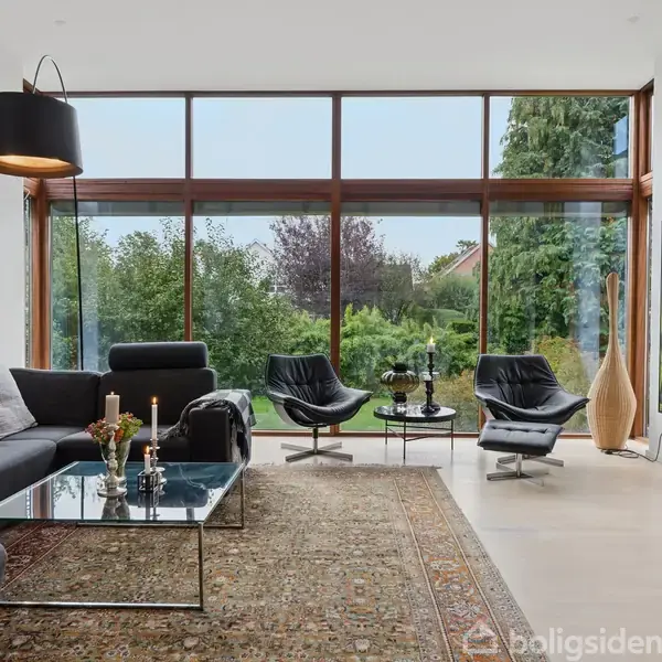 Stue med stor glasvæg giver udsigt til grøn have. Moderne sofa og lænestole centreret omkring et glasbord med stearinlys. Væggene prydet med farverige kunstværker.