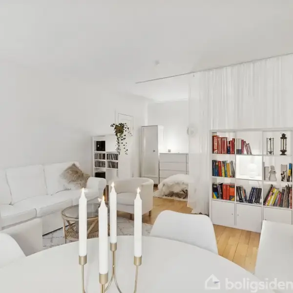 En stue med hvid sofa, sofabord og puder arrangeret rundt om et rundt bord med stearinlys. En bogreol med bøger og dekorative genstande er i baggrunden. Rummet er lyst og minimalistisk.