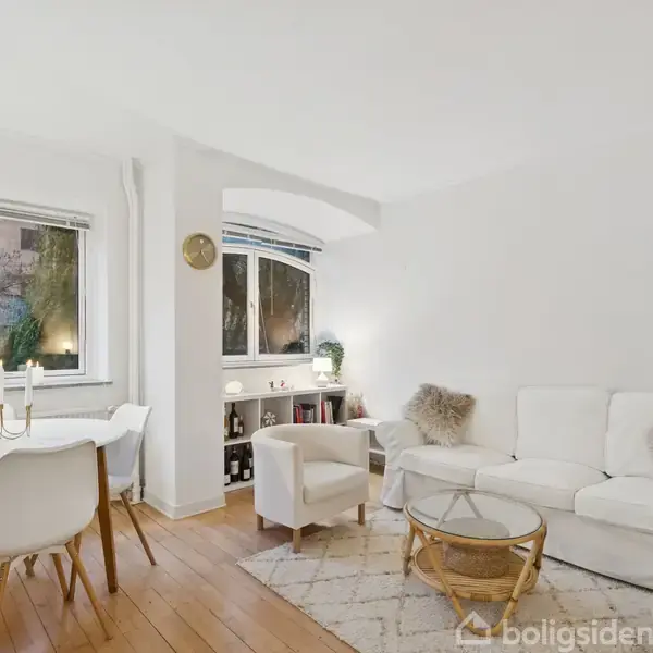 En lys stue med en hvid sofa og et rundt sofabord står på et trægulv. En spiseplads med fire stole ved et rundt bord er ved siden af store vinduer.