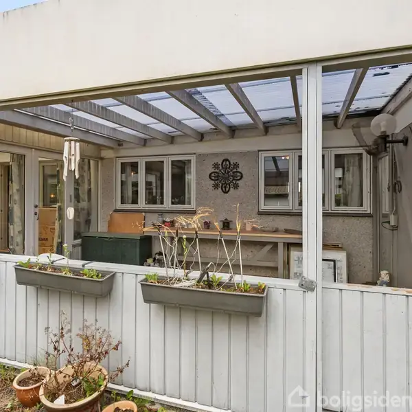 Terrasse indrettet med plantekasser, borde og stole under et halvtag. Vinduerne omkranser området, og der hænger en lanterne og dekoration på væggen. Grønne planter og blomster er til stede.