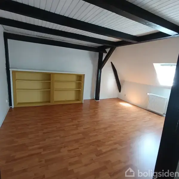 Reol står mod en hvid væg uden bøger. Den er i et rum med trægulv, loftsbjælker og et skråvindue, der lader lys ind.