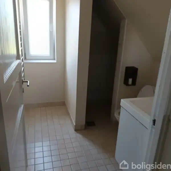 Badeværelse med klinkegulv. Toilet ved siden af en hvid vask. Smalt vindue til venstre. Dusjområde i baggrunden til højre. Vægge i neutrale farver.