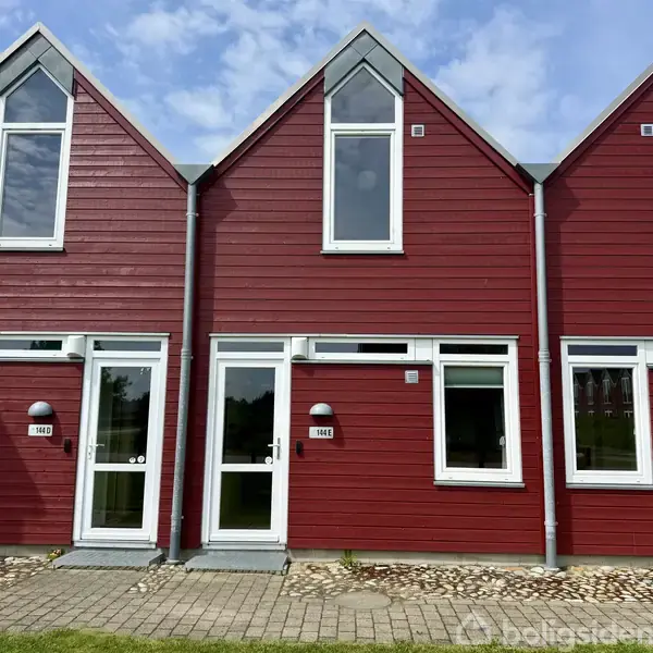Rødt rækkehus med hvidrammede vinduer og døre står på en flisebelagt forplads. Tekst på skilt ved dør: "184E". Omkringliggende huse har samme stil under en blå, skyet himmel.