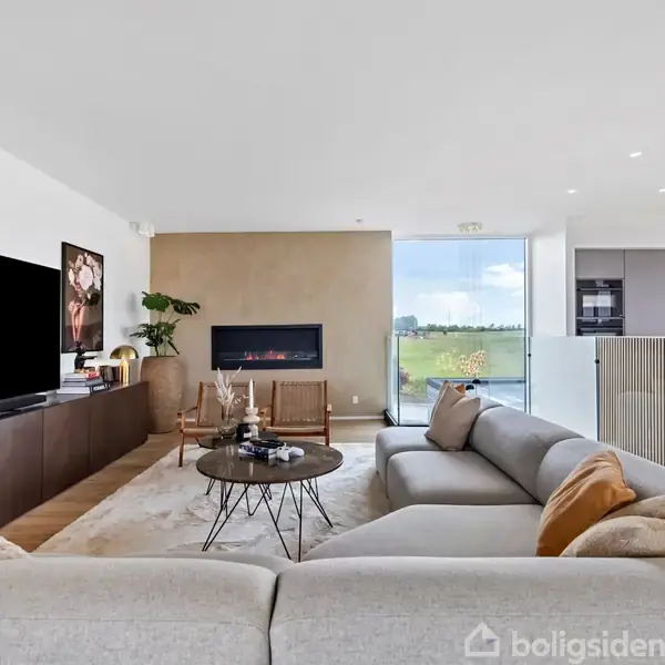 Stor, grå sofa står i en moderne stue med et lavt sofabord. Til venstre ses et stort TV over en skænk. Bagved er en vægpejs foran en stor glasdør med udsigt.