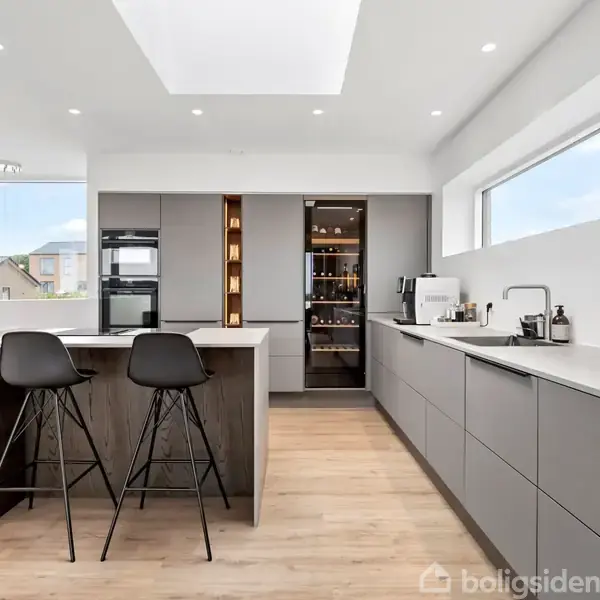 To høje barstole står ved en køkkenø, omgivet af et moderne køkken med grå skabe. Store vinduer til højre og et lyst loft skaber en rummelig atmosfære.