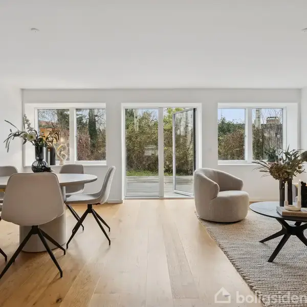 Spisestue med rundbord og stole, placeret i et lyst, moderne værelse med trægulv. Store vinduer og glasdøre fører ud til en have. Indretningen er minimalistisk.