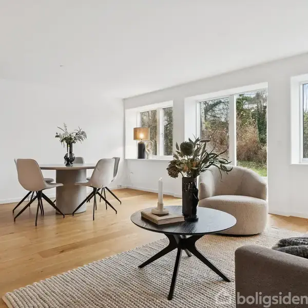 Et moderne opholdsrum med trægulv, hvor en sofa og et rundt sofabord med dekorative planter står. Et spisebord med fire stole er placeret ved vinduer, der viser en skovudsigt.