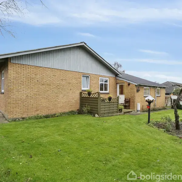 Et enfamiliehus i gule mursten står på en grøn græsplæne. Huset har en lille terrasse med rækværk, omgivet af en have med buske og træer under en blå himmel.