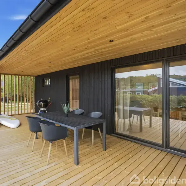 Terrasse med træbord og fire stole står under træoverdækning. Glidende glasdøre åbner ind til huset. Et grill står i baggrunden, omkranset af natur og huse i afstanden.