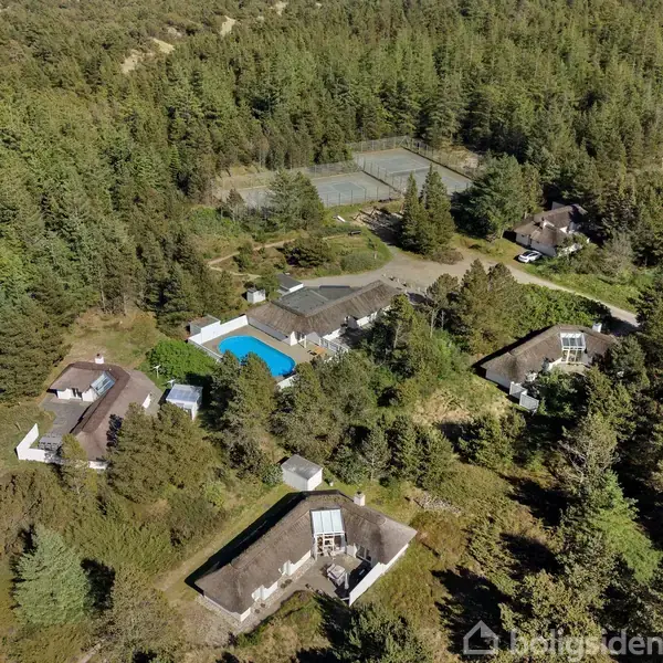 Et kompleks med flere huse omgivet af frodig skov. Et centralt hus har en klarblå rektangulær swimmingpool ved siden af. Området ligger i en tæt, skovklædt landskab.