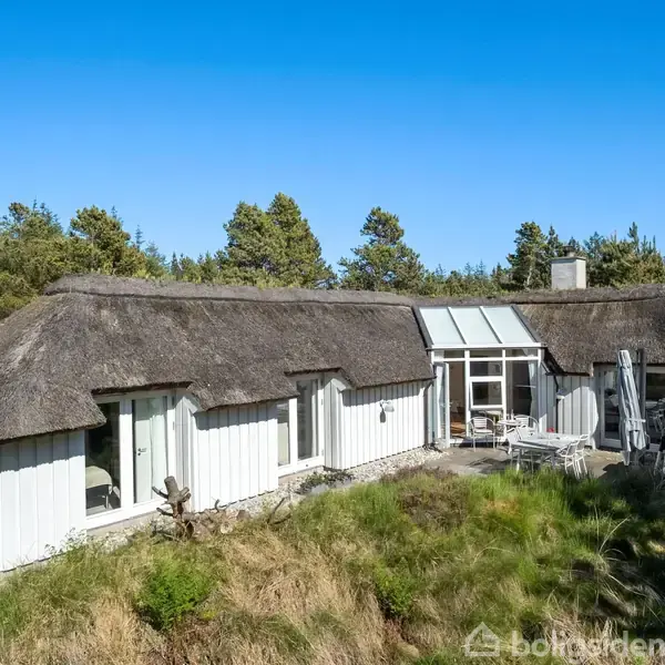 Et stråtækt hus med hvide vægge står i en grøn, skovklædt omgivelser. Sollys skinner på terrassen med havemøbler foran store vinduer.