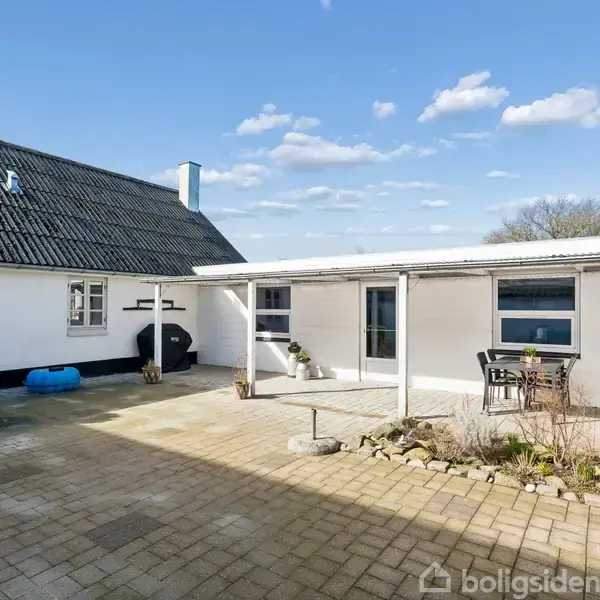 Hvid hus omgiver flisebelagt gård, der rummer bord og stole under overdækket terrasse. Grønne planter pryder området. Klar, blå himmel skaber lys atmosfære.