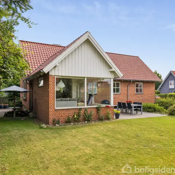 Et rødt murstenshus med rødt tegltag står i en grøn have med en terrasse til højre. Udendørsmøbler og en parasol er placeret ved siden af huset.